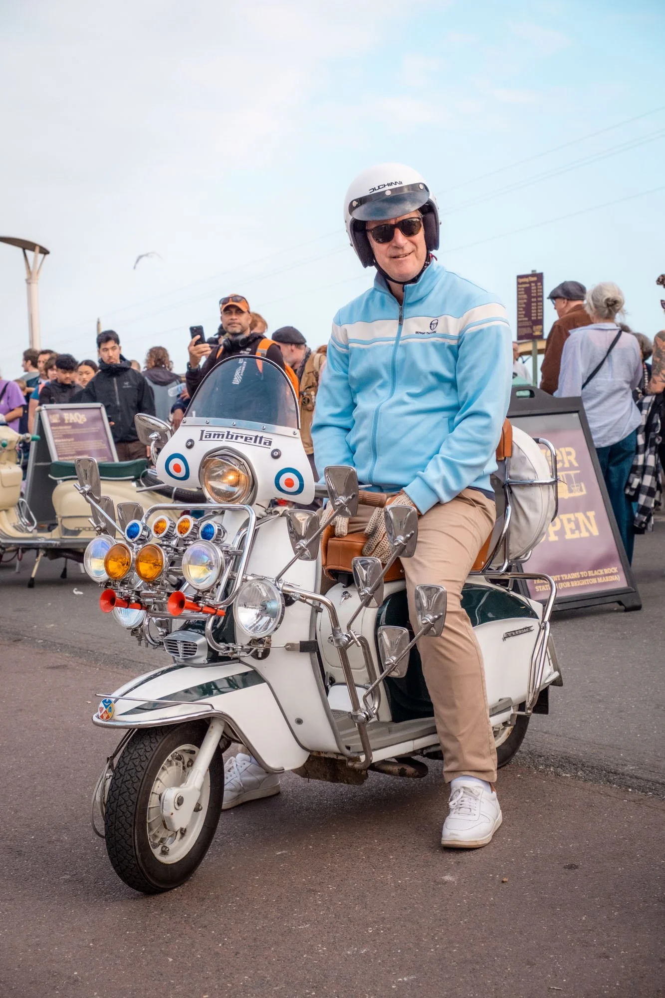 Man wearing sunglasses and a helmet sitting on a white vintage scooter at a crowded outdoor event.