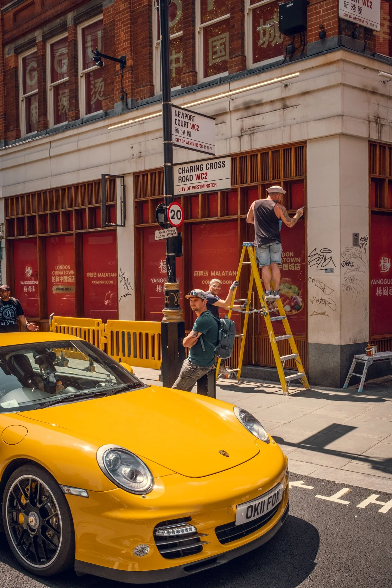 A bright yellow Porsche parked on the street with a building and street signs behind it. Two men, one on a ladder and another standing next to him, appear to be working on something at the corner of the building. A woman is walking past, and another 