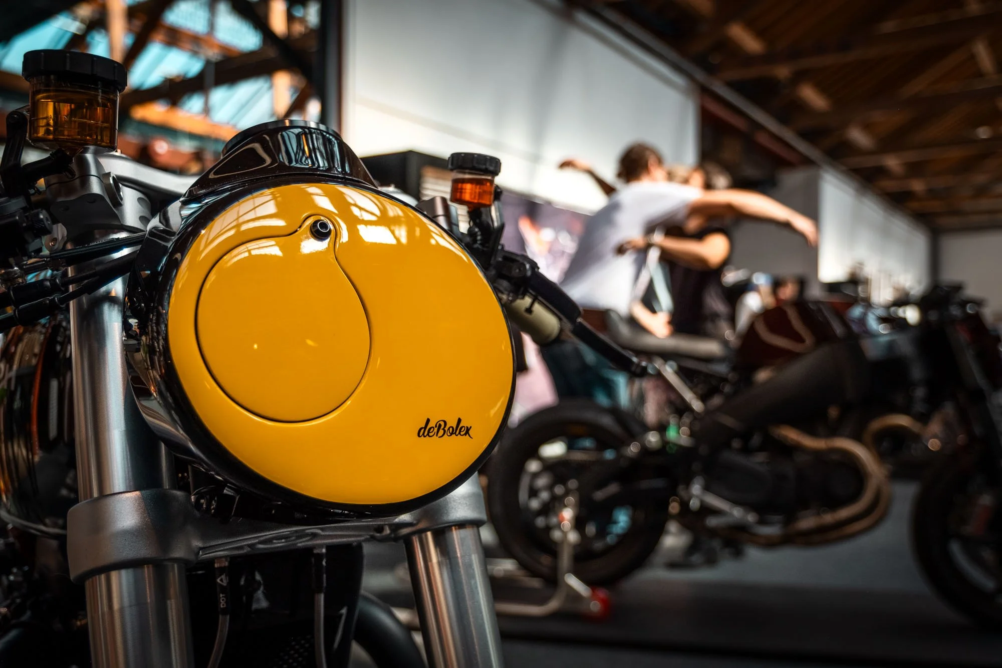 Close-up of a yellow motorcycle gas tank with the brand name 'deBolex' in black, in a showroom with people and other motorcycles in the background.