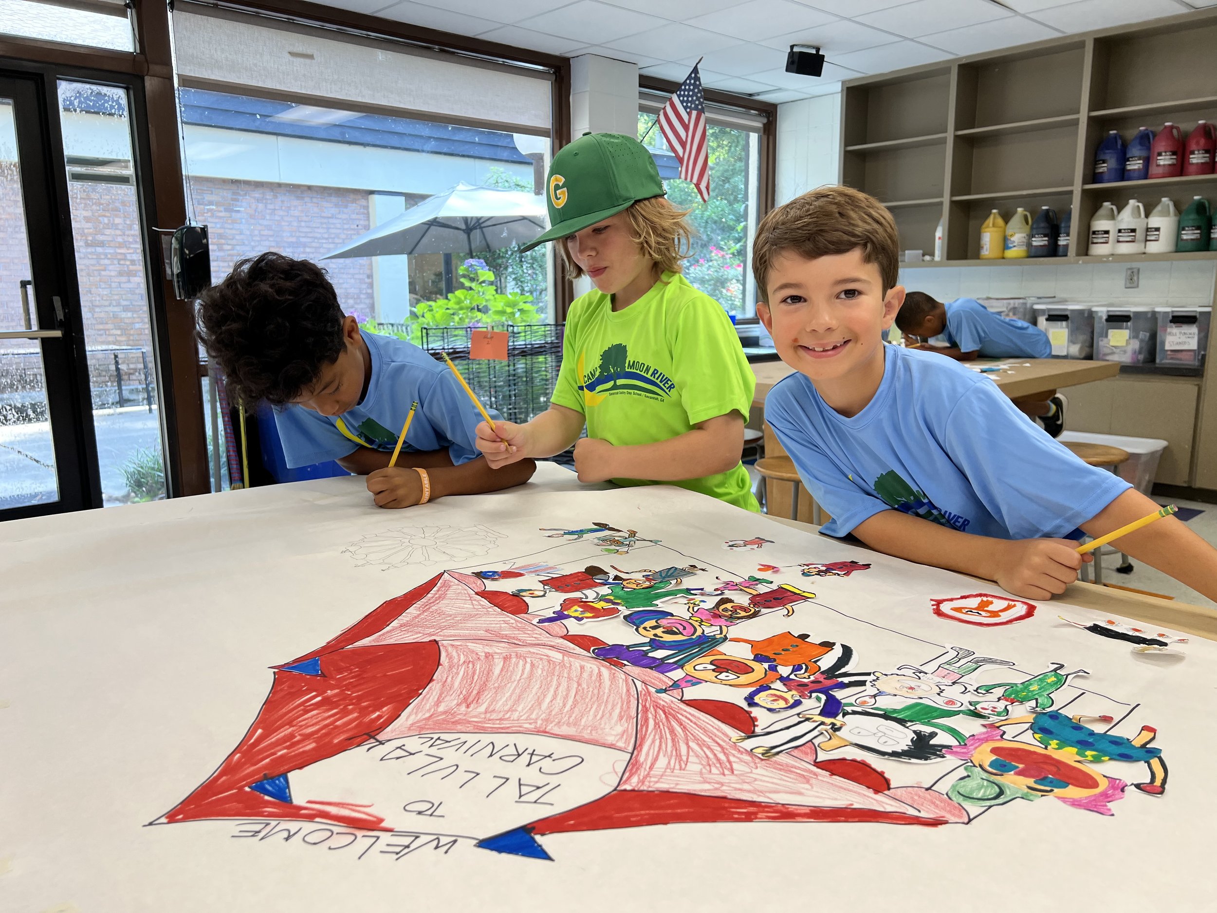 Three children drawing on a large paper with colorful illustrations, inside a classroom with art supplies and an American flag in the background. They are engaged in creating a "Welcome to Talulah Carnival" poster.