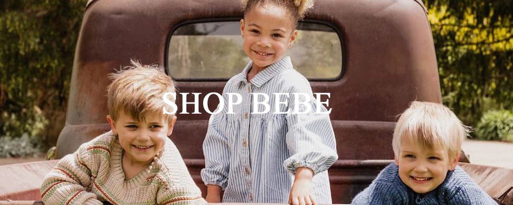 Three young children sitting on the back of a vintage pickup truck outdoors, smiling at the camera. Text overlay reads, "SHOP BEBE." The scene is bright and cheerful.