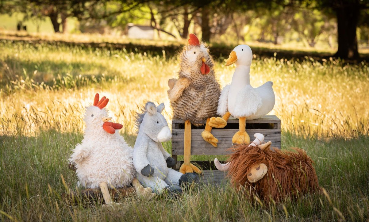 Multiple plush animal puppets, including a chicken, horse, duck, cow, and lion, arranged outdoors on grass with trees in the background.