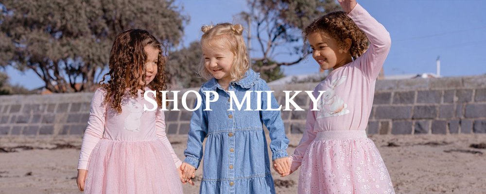Three young girls holding hands and smiling on a beach, with a stone wall and trees in the background, wearing casual dresses.