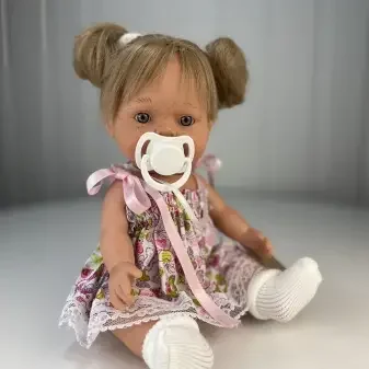 Young girl with pigtails wearing a floral dress and white shoes, sitting on the floor with a pacifier in her mouth.