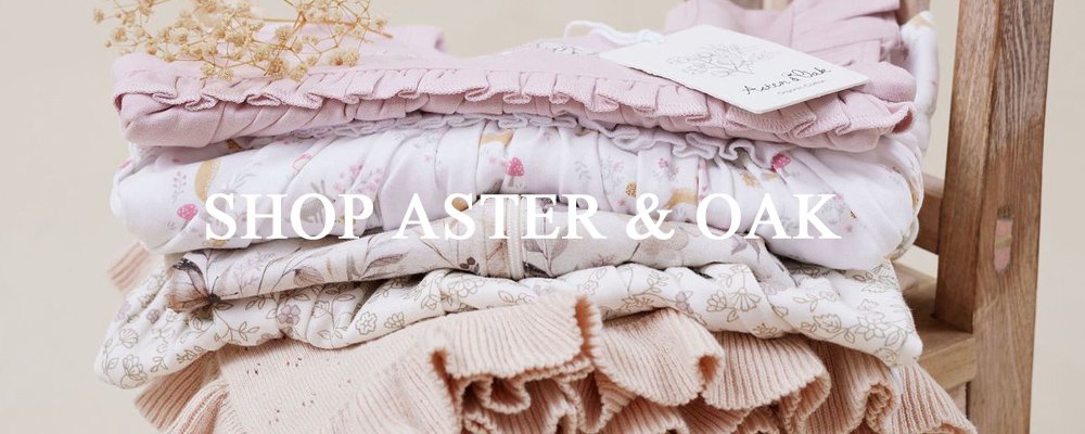 Stacked folded pastel-colored clothing including pink, white, and peach, with a floral or abstract pattern, on a wooden surface with a light pink blanket or towel on top; text overlay reads 'SHOP ASTER & OAK'.