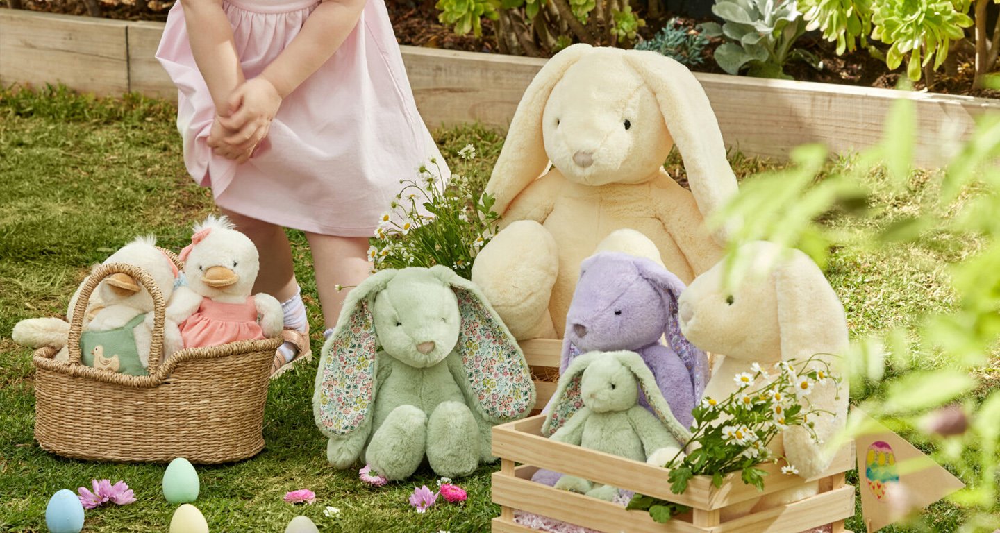 A young girl standing outdoors near a display of plush bunny and duck toys, some in baskets and small wooden crates, surrounded by flowers and grass.