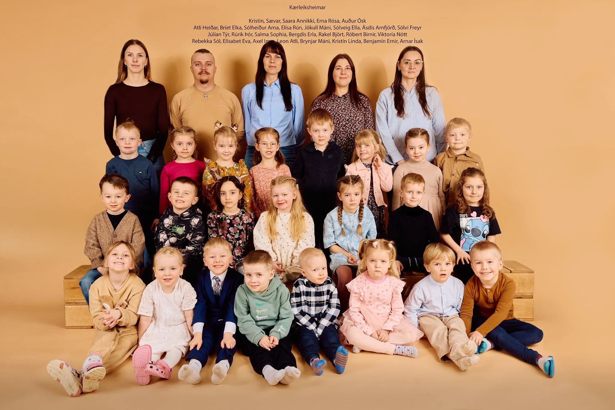A group photo of children and teachers standing and sitting against a beige background, with names listed at the top.
