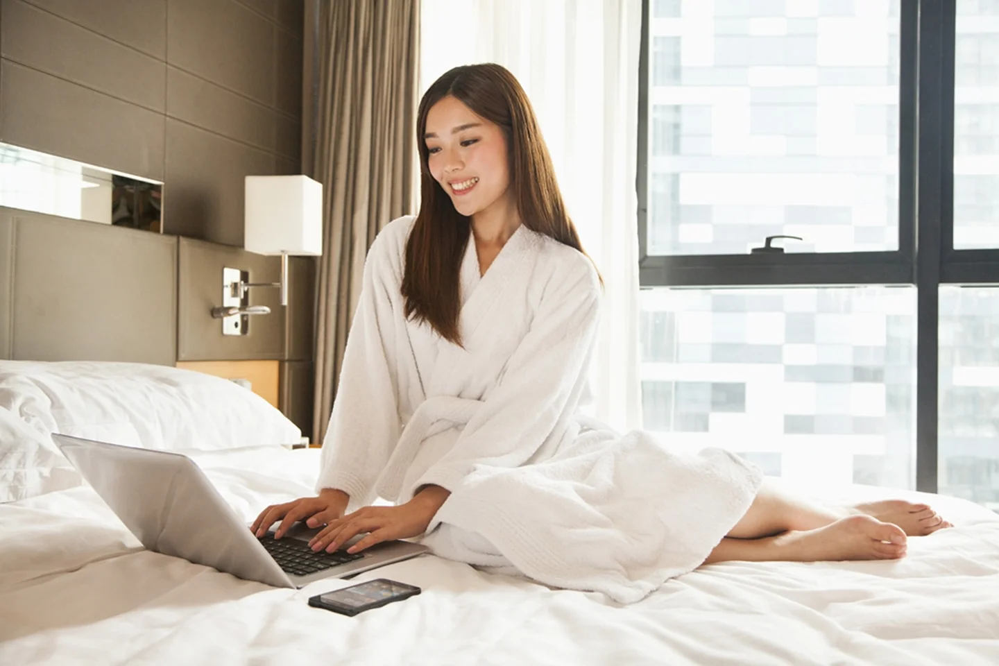 Beautiful Woman in Robe Sitting on Bed Smiling and Using Laptop