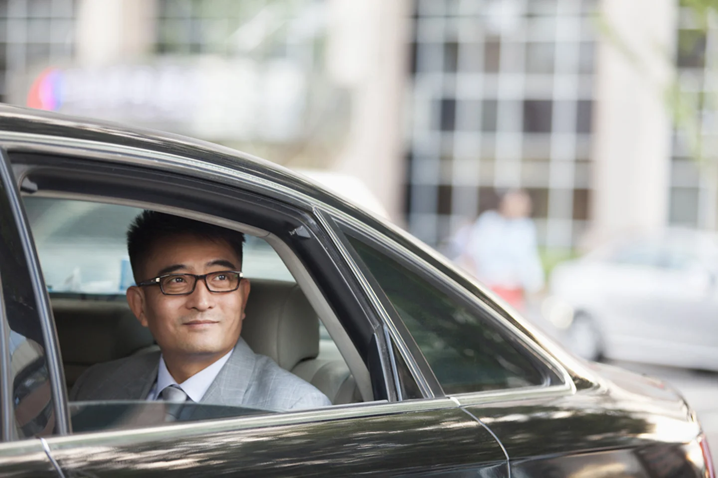 Man in business attire with glasses peering out from backseat of car
