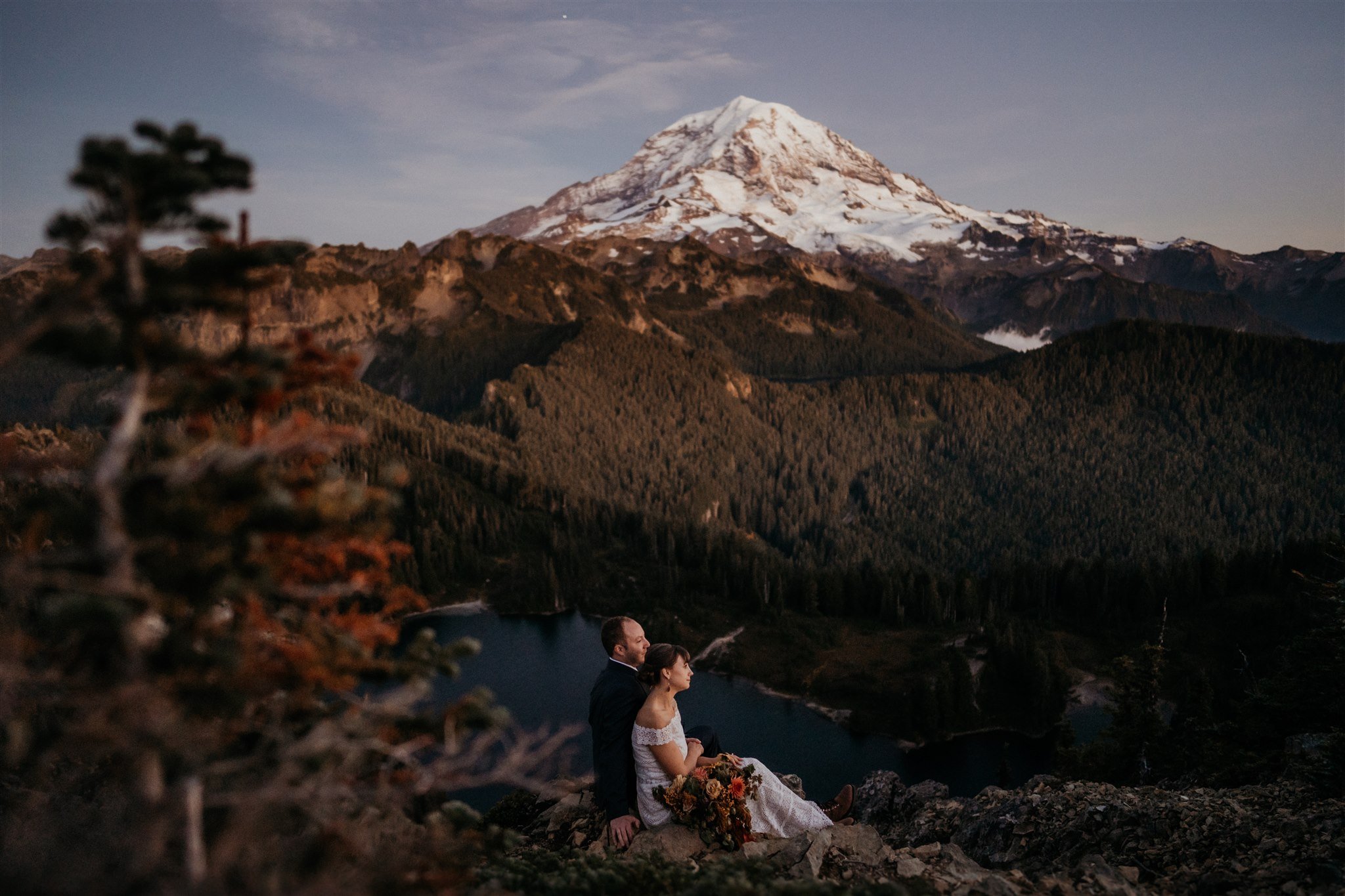 Bride and groom sit on a ledge overlooking the lake and Mount Rainier