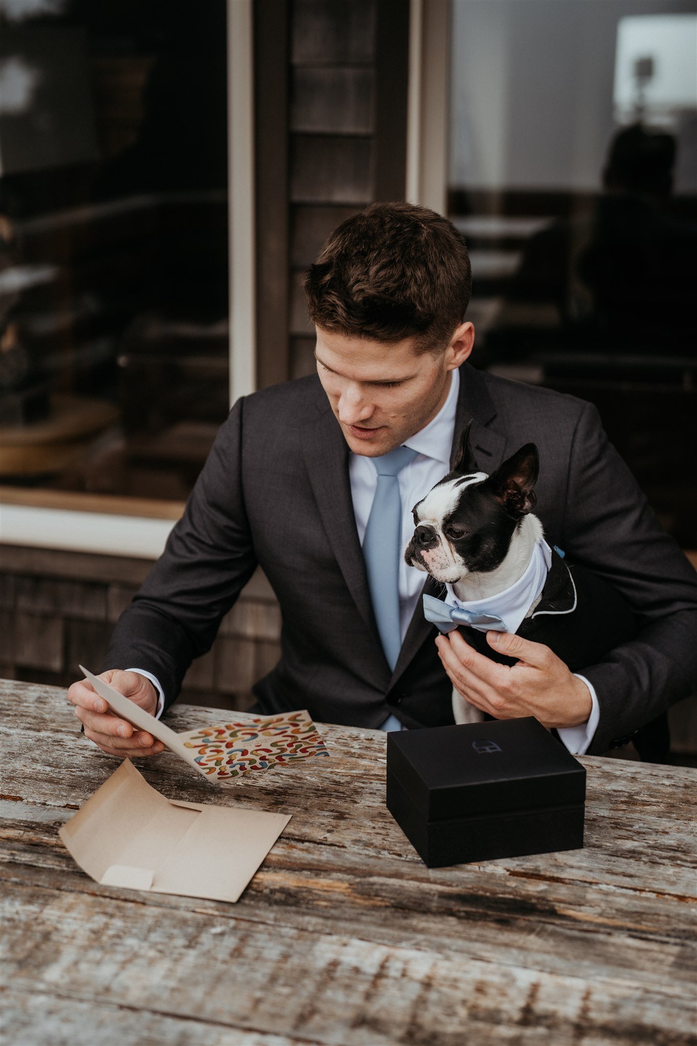 Groom holding pug dog while reading handwritten card at a picnic table