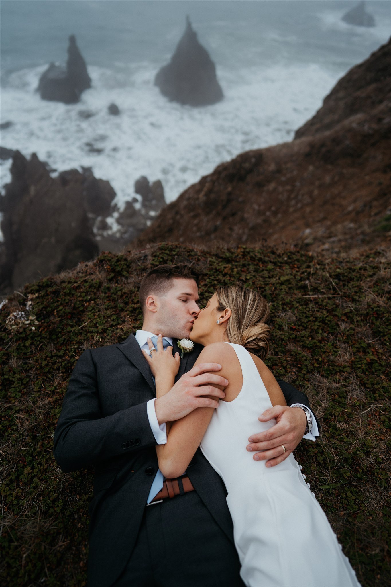 Bride and groom laying down on the ground and kissing by the cliff on the Southern Oregon Coast