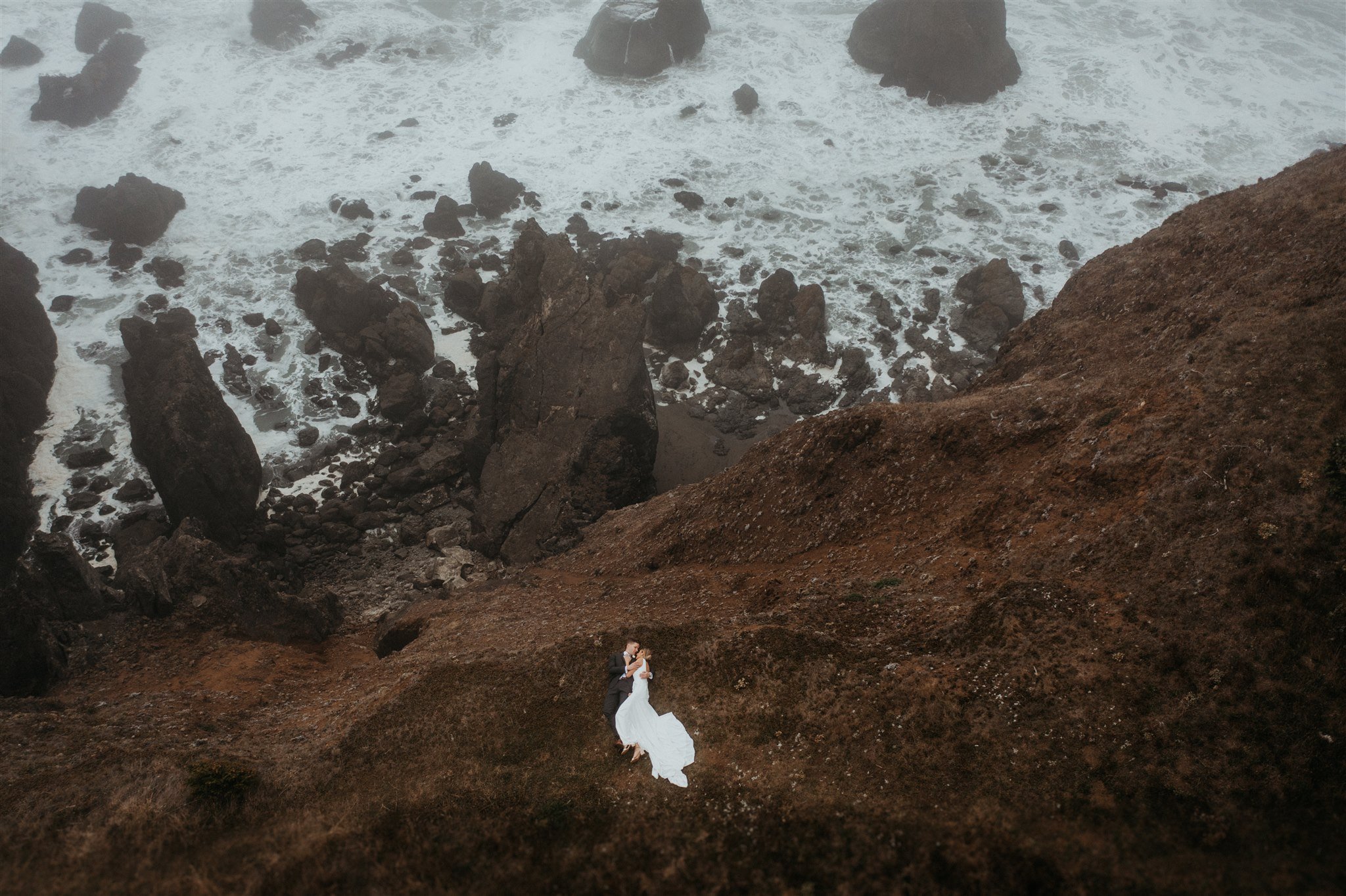 Bride and groom laying down on the ground and kissing by the cliff on the Southern Oregon Coast