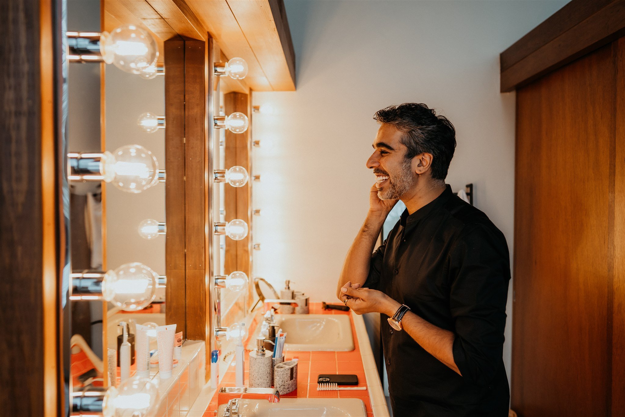 Groom smiling and getting ready in front of lightbulb mirror in Colorado mountain cabin