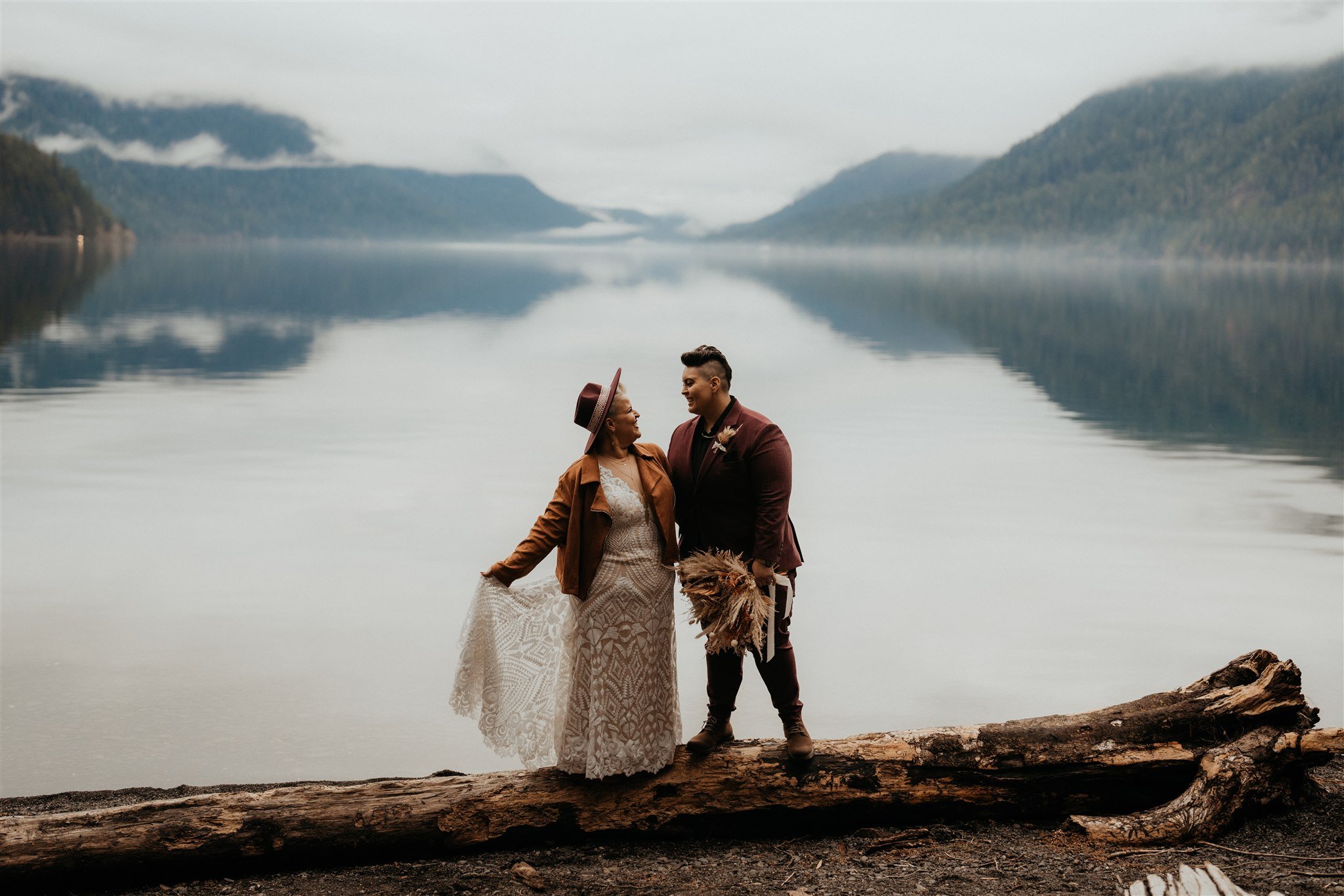 Two brides standing on a fallen log in front of a lake in La Push