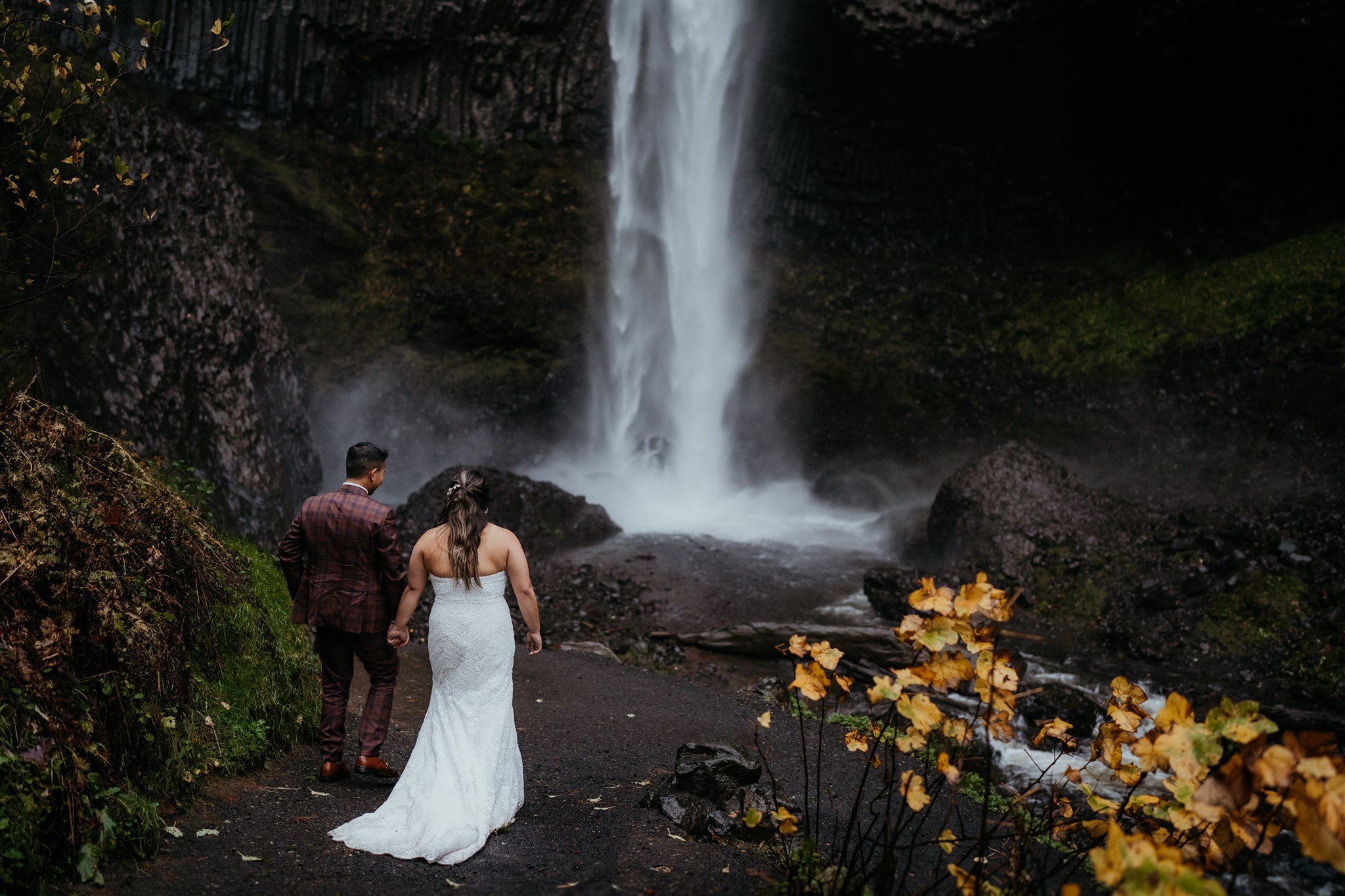 Bride and groom hold hands while walking down a trail towards a waterfall