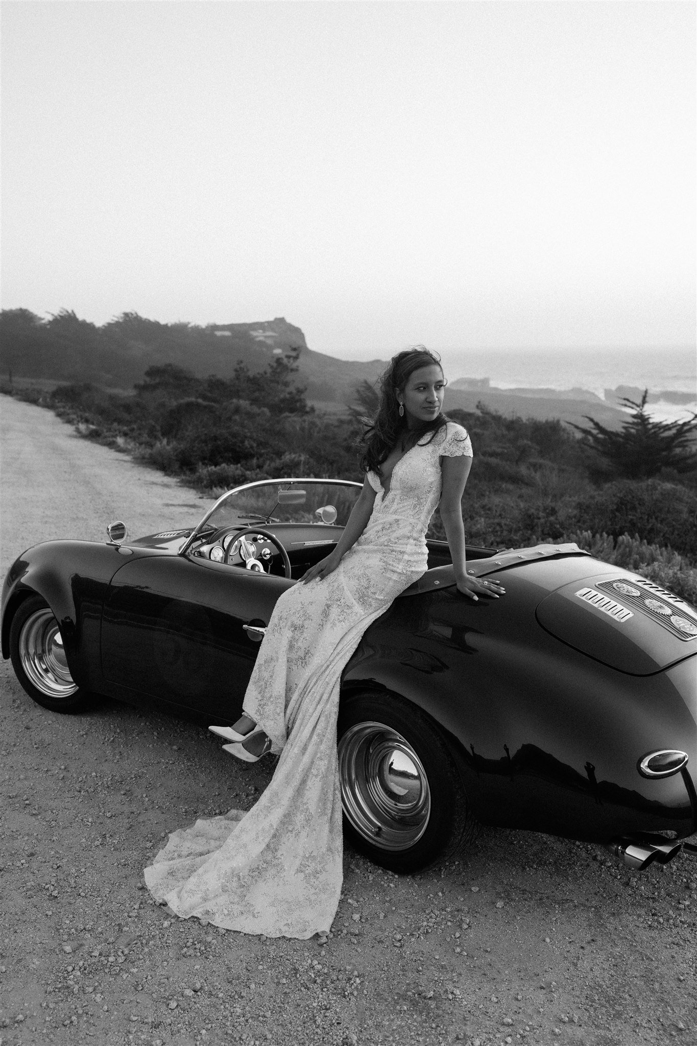 Bride leaning against a vintage convertible car on the side of the road in Big Sur