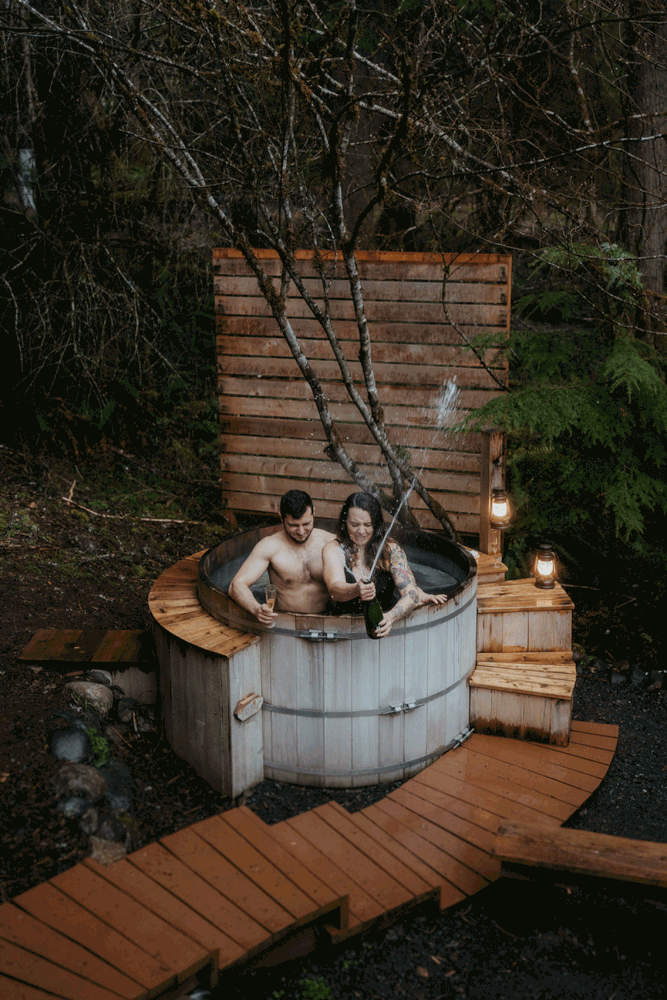 Bride and groom spray champagne in barrel hot tub after snowy hiking elopement