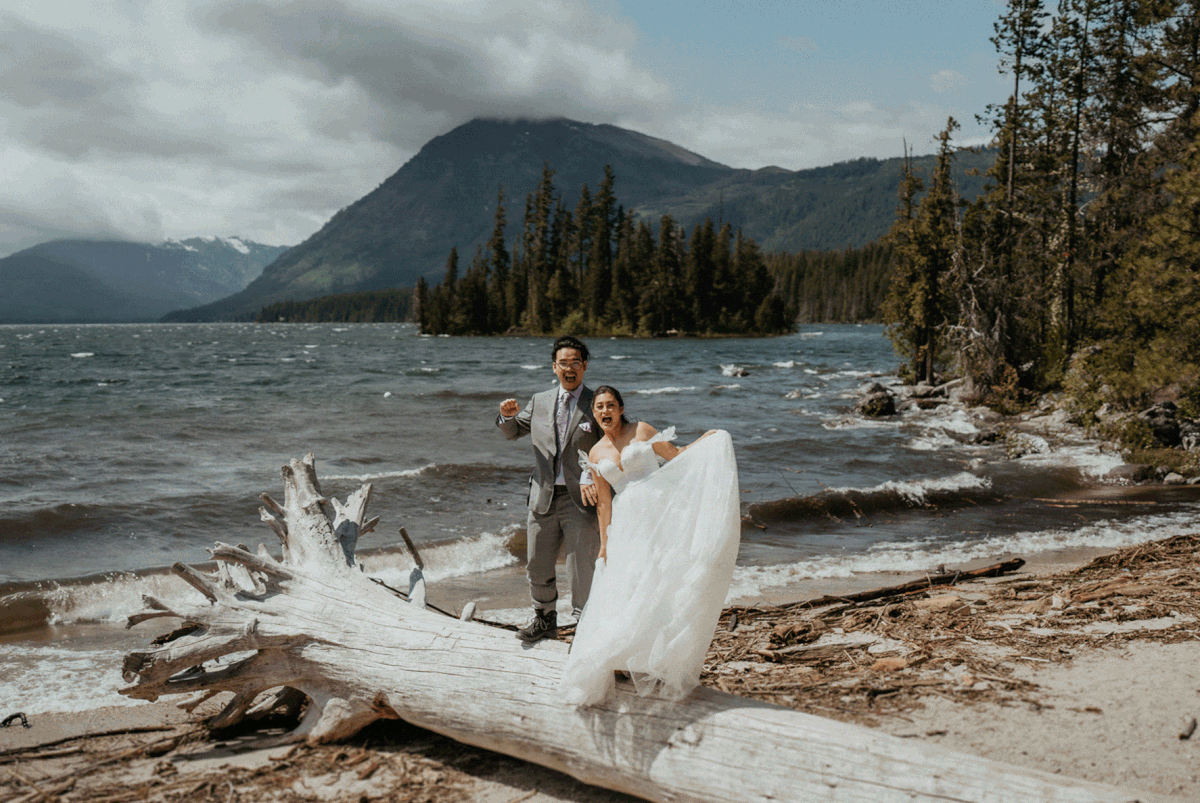 Bride and groom cheer while standing on a tree stump by the lake