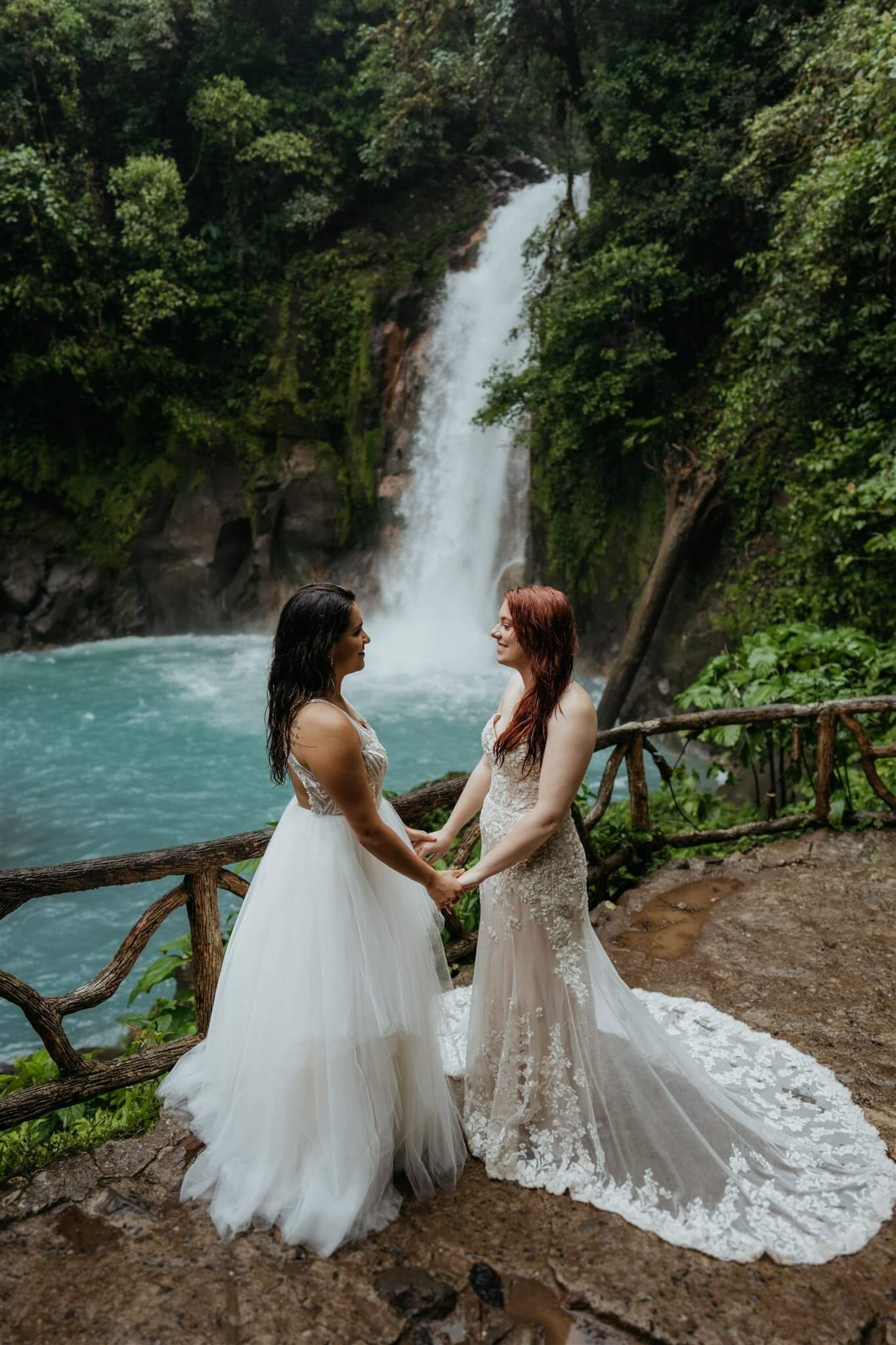 Two brides hold hands next to a waterfall during destination wedding in Costa Rica