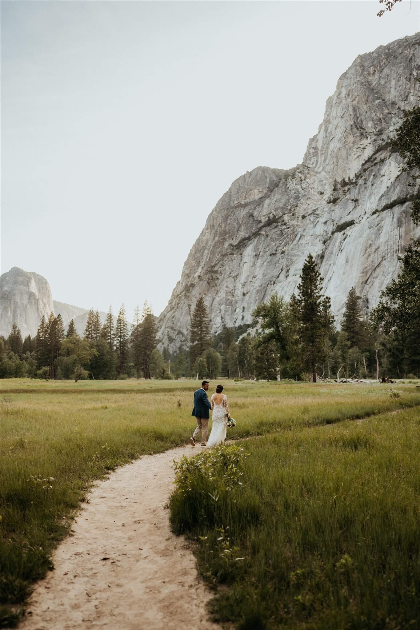 Bride and groom hold hands and talk while walking across the meadow at Yosemite