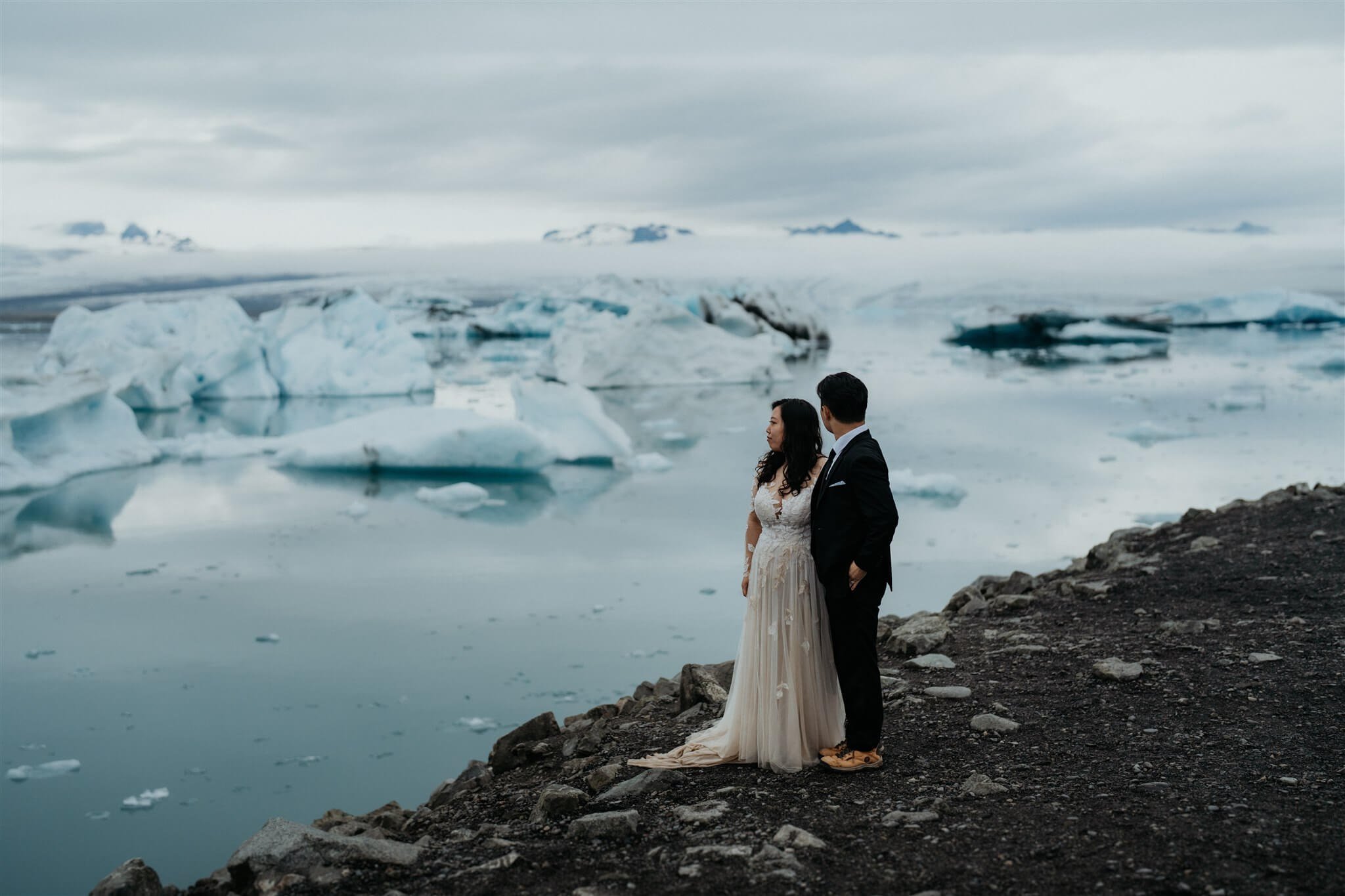 Bride and groom standing on a black sand beach by a lagoon in Iceland