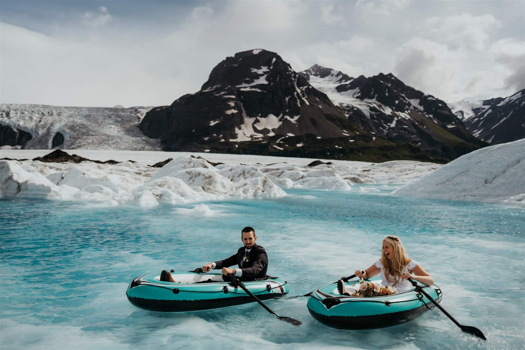 Bride and groom paddle inflatable boats on a glacier pond in Alaska