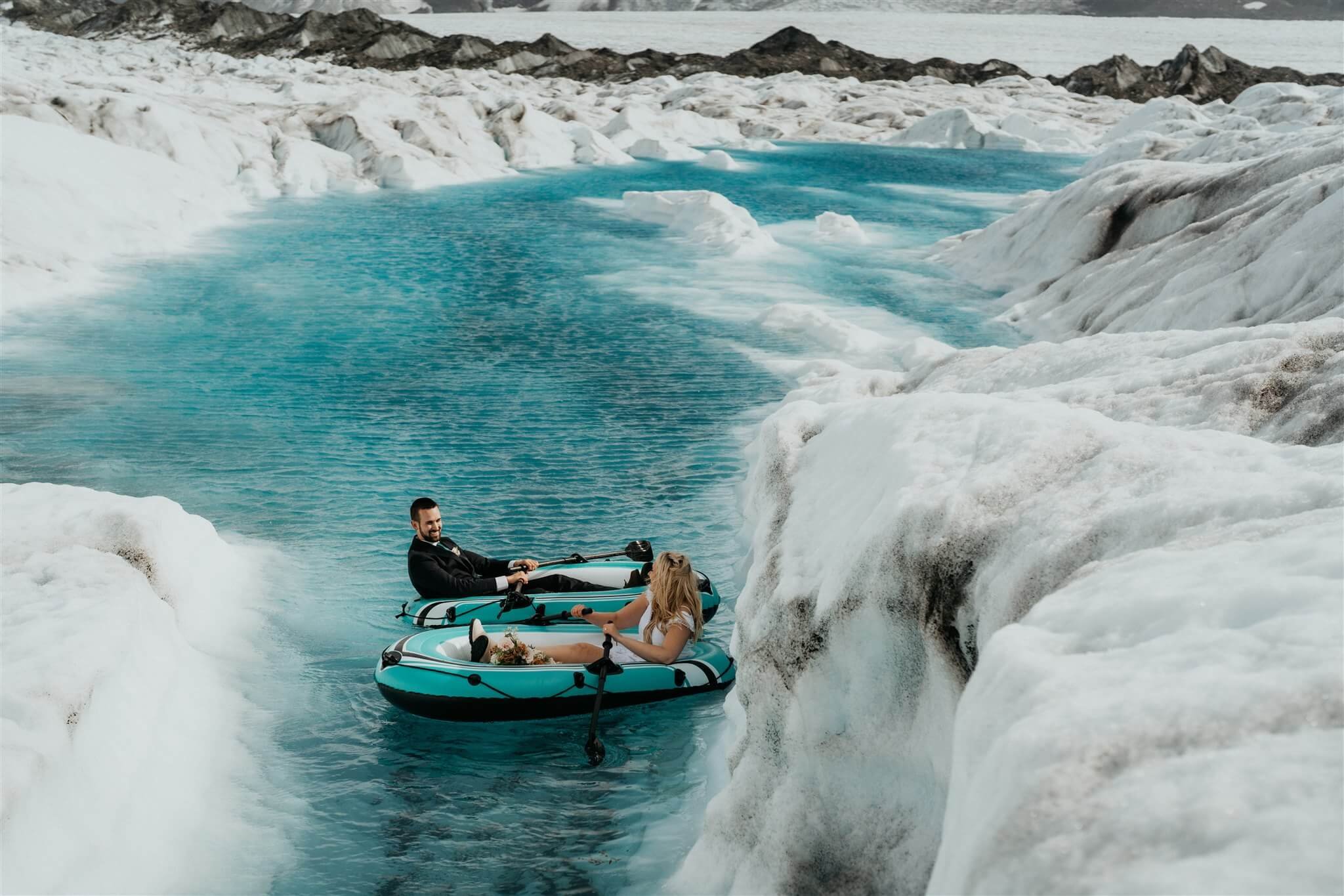 Bride and groom paddle inflatable boats on a glacier pond in Alaska