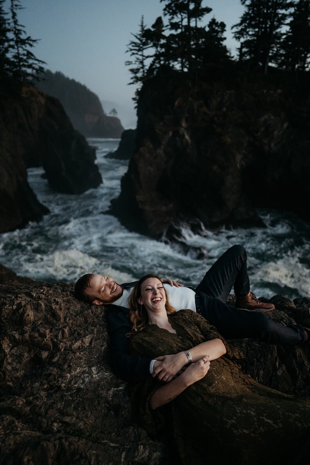Couple laughing while laying down on the cliffs at their Oregon Coast couple photo shoot