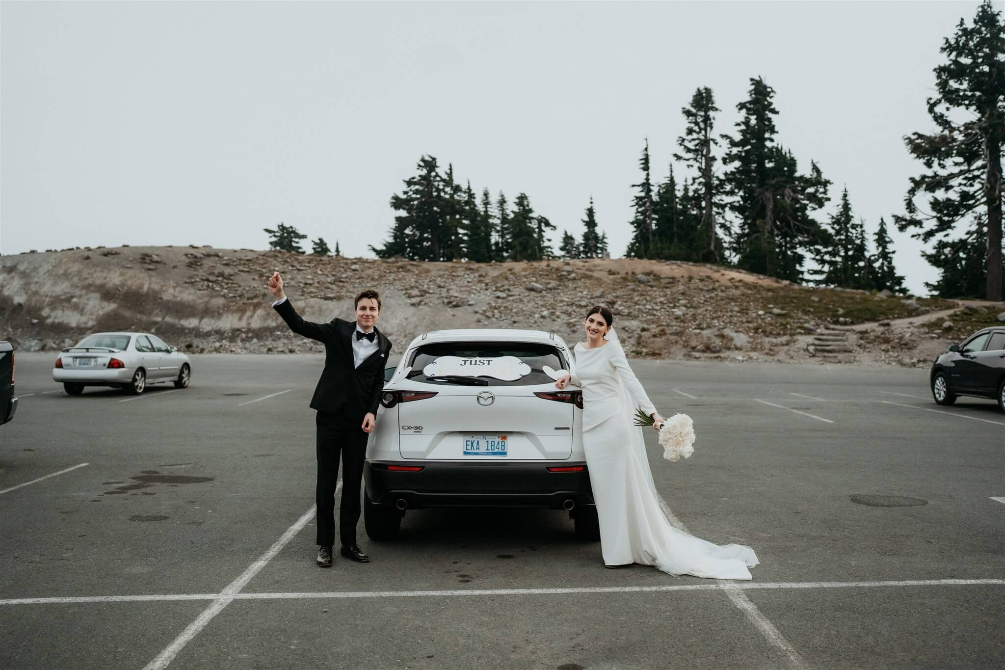 Bride and groom standing next to their white car with a "Just Married" sign on the back window