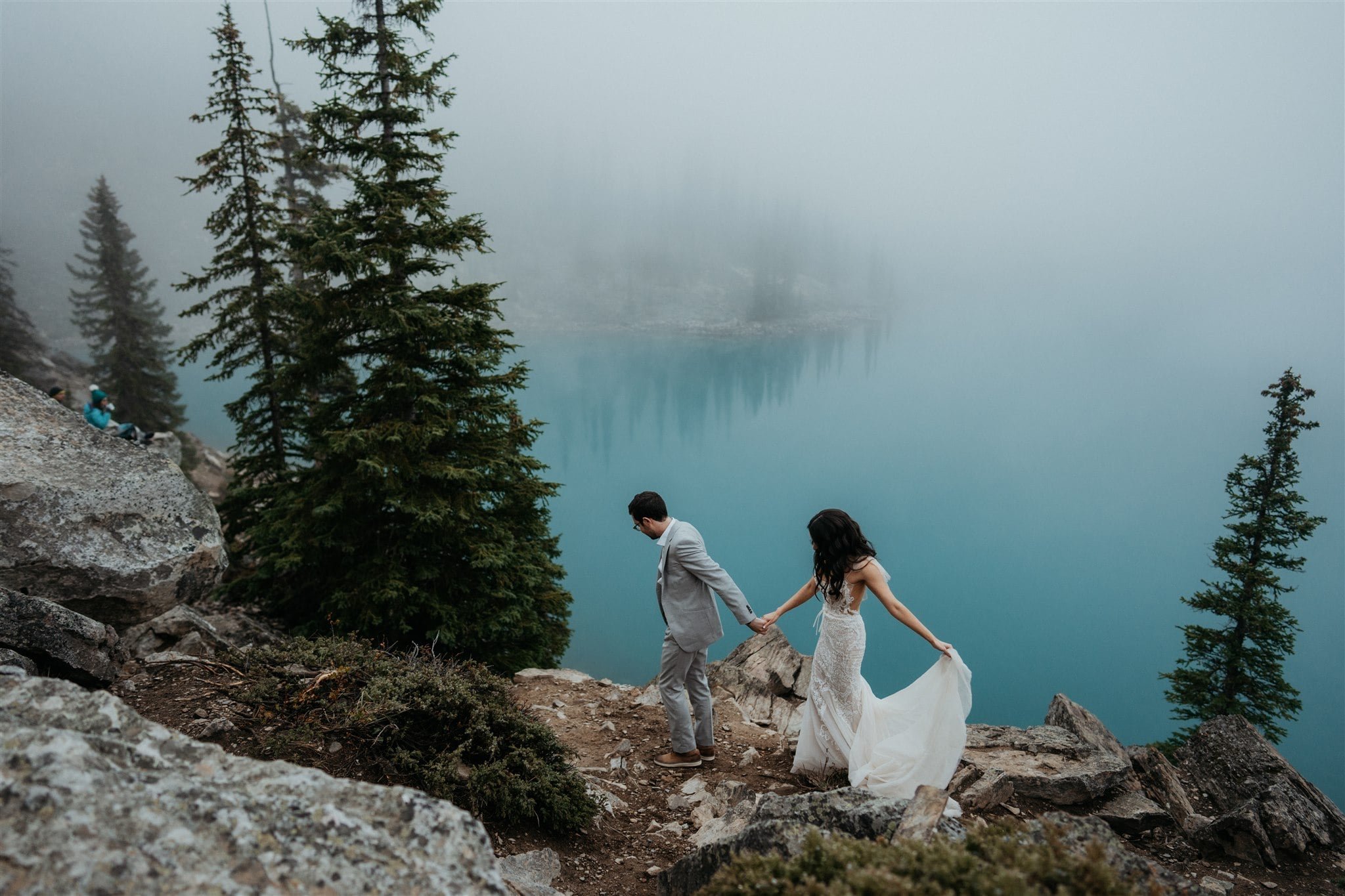 Bride and groom hold hands while walking across a mountain trail at their Banff elopement