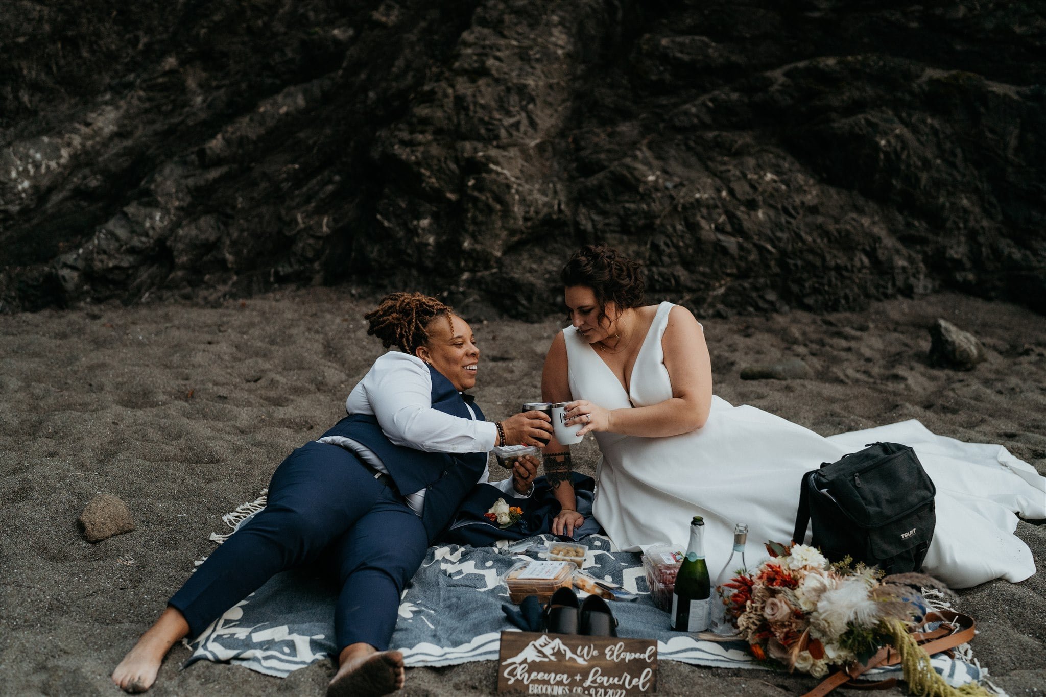 Two brides lounge on a picnic blanket on the beach during their Southern Oregon Coast elopement