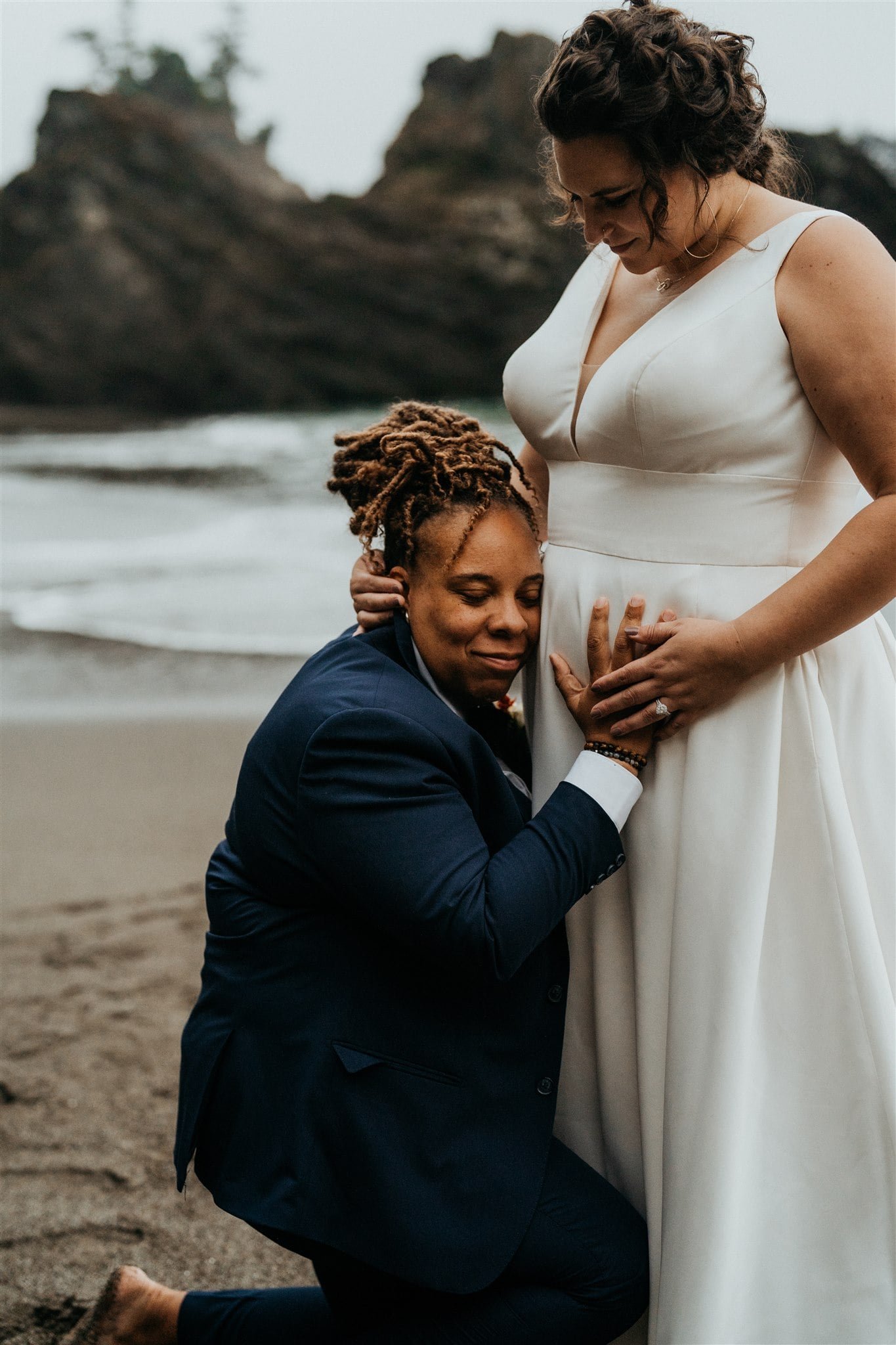 Bride rests her head on her wife's belly during their beach elopement on the Oregon Coast
