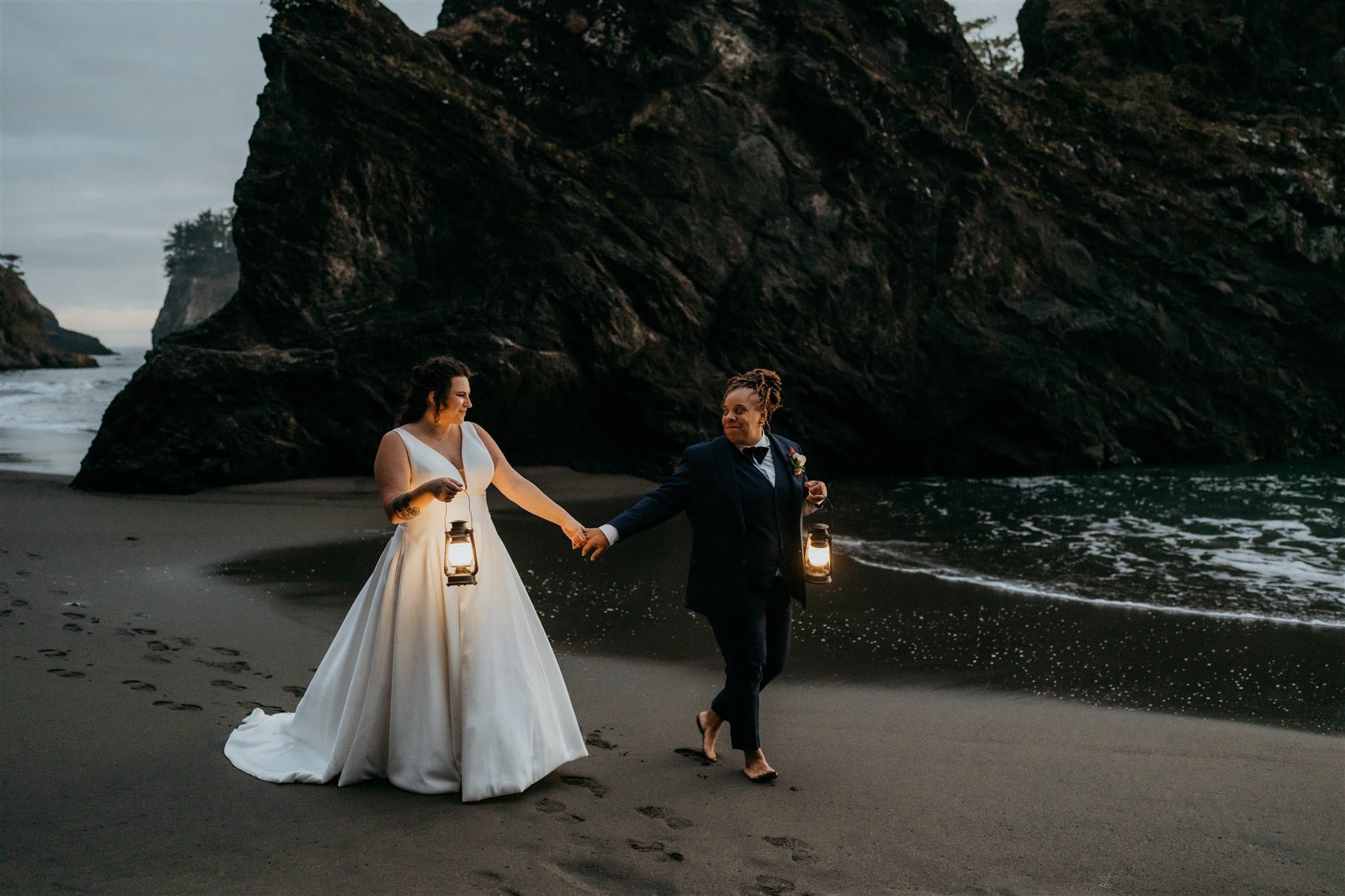 Two brides holding lanterns and walking along the beach on the Oregon Coast during sunset