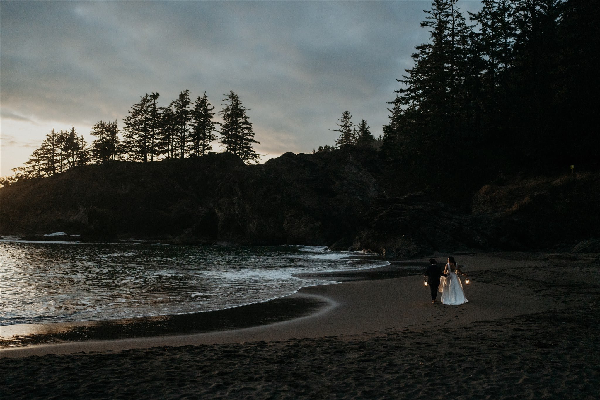 Two brides holding lanterns and walking along the beach during their Oregon Coast elopement