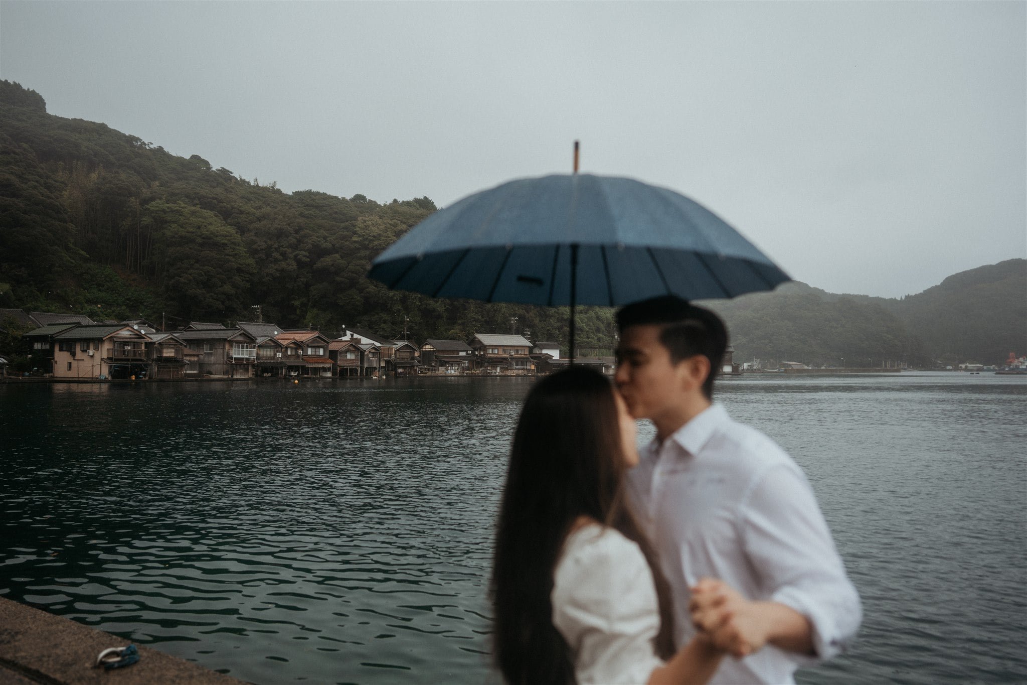 Groom kissing bride on the forehead while standing under a blue umbrella