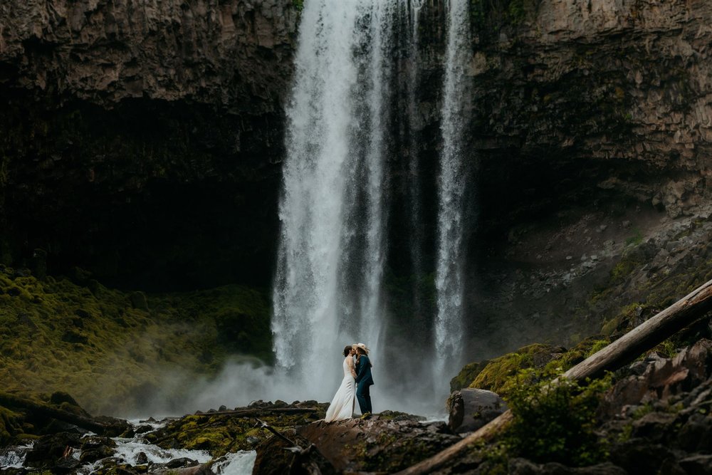 Two brides kiss in front of a waterfall during their Oregon elopement