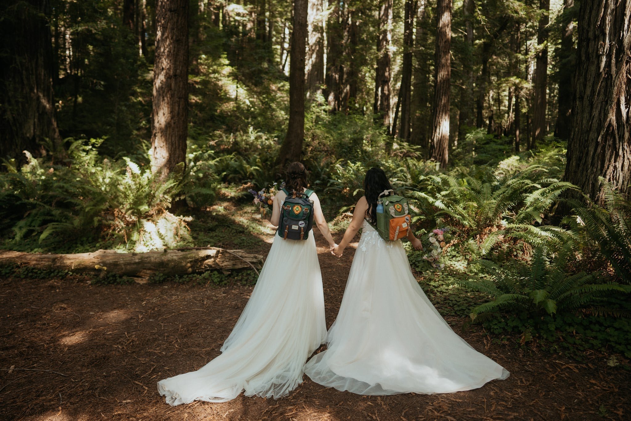 Two brides hiking through the woods while wearing matching backpacks for their elopement at Secret Beach, Oregon