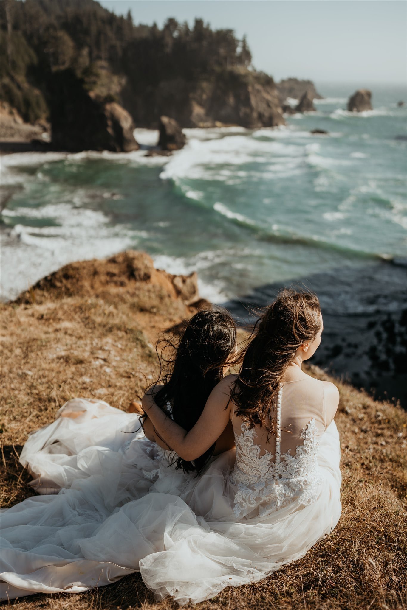 Two brides sitting on a cliff overlooking the ocean during their Secret Beach, Oregon elopement
