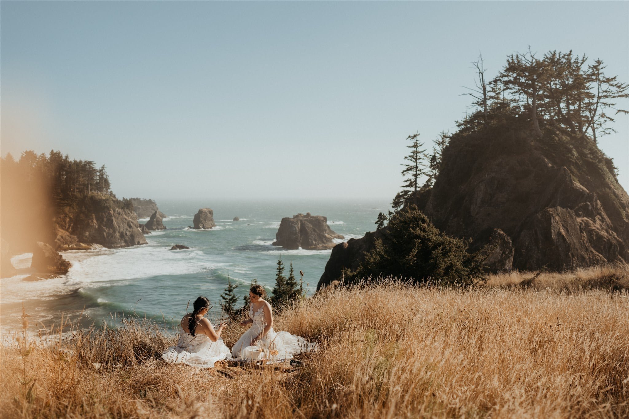 Two brides cutting cake while sitting on a cliff overlooking the ocean during their Secret Beach, Oregon elopement