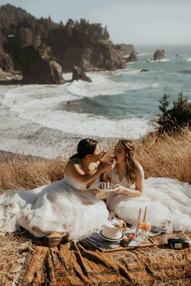 Two brides eating cake while sitting on a cliff overlooking the ocean during their Secret Beach, Oregon elopement