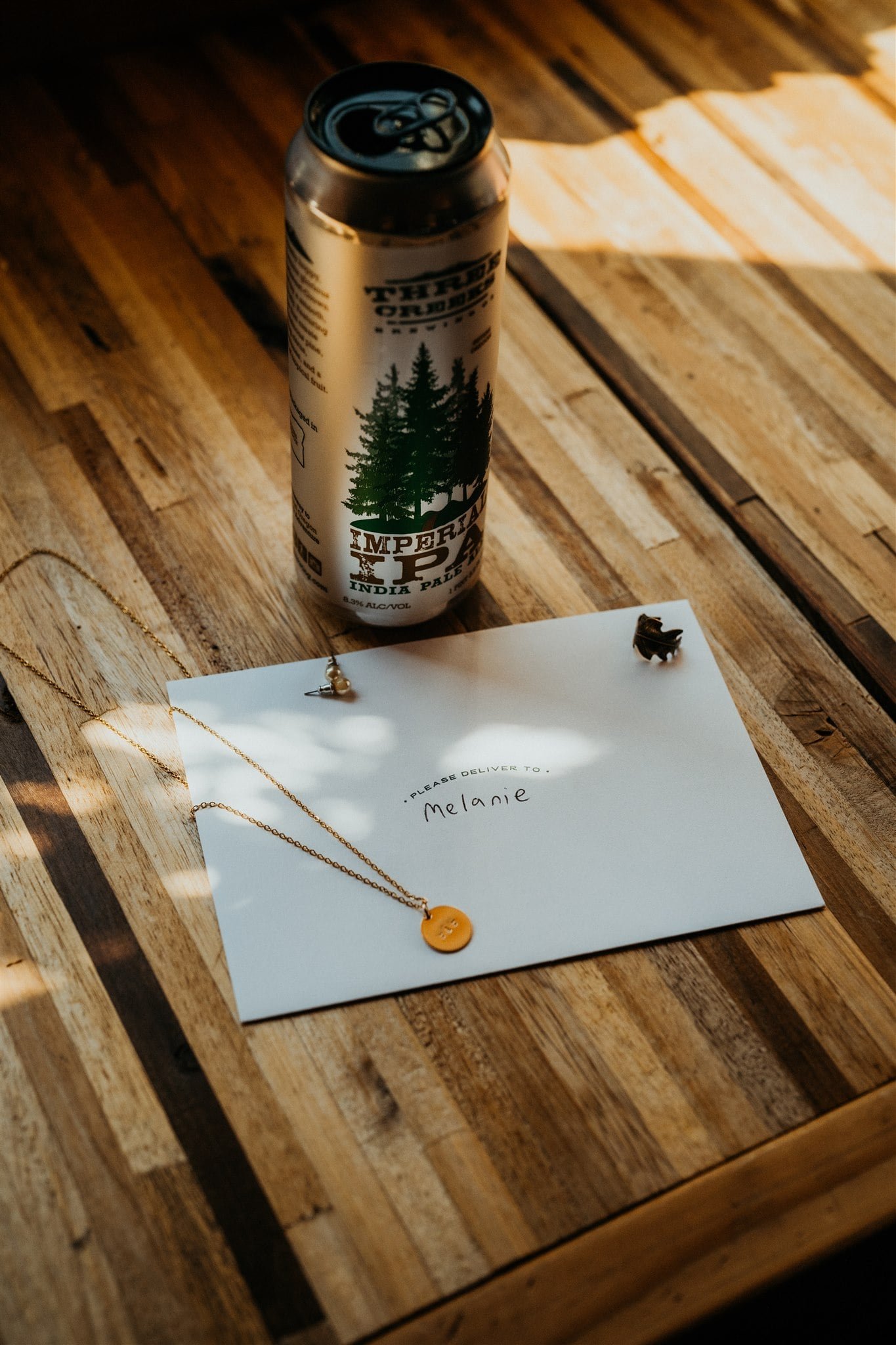 White envelope that reads "please deliver to Melanie" with IPA beer and gold necklace