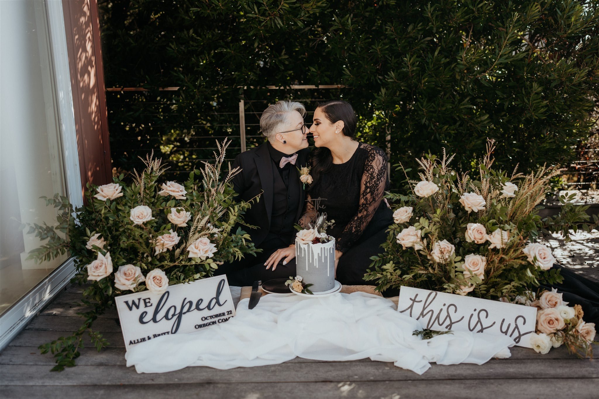 Two brides sit on the ground with a gray frosted elopement cake and neutral wedding flower arrangements