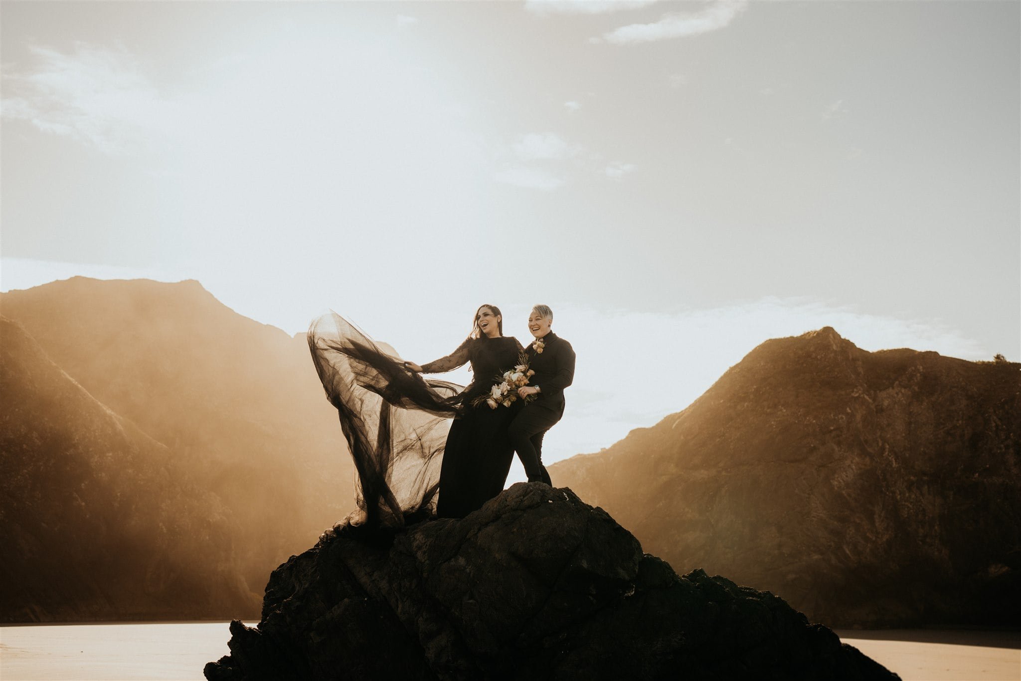 Two brides wearing all black wedding attire standing on a rock on the Oregon Coast for their Pacific Northwest elopement photos