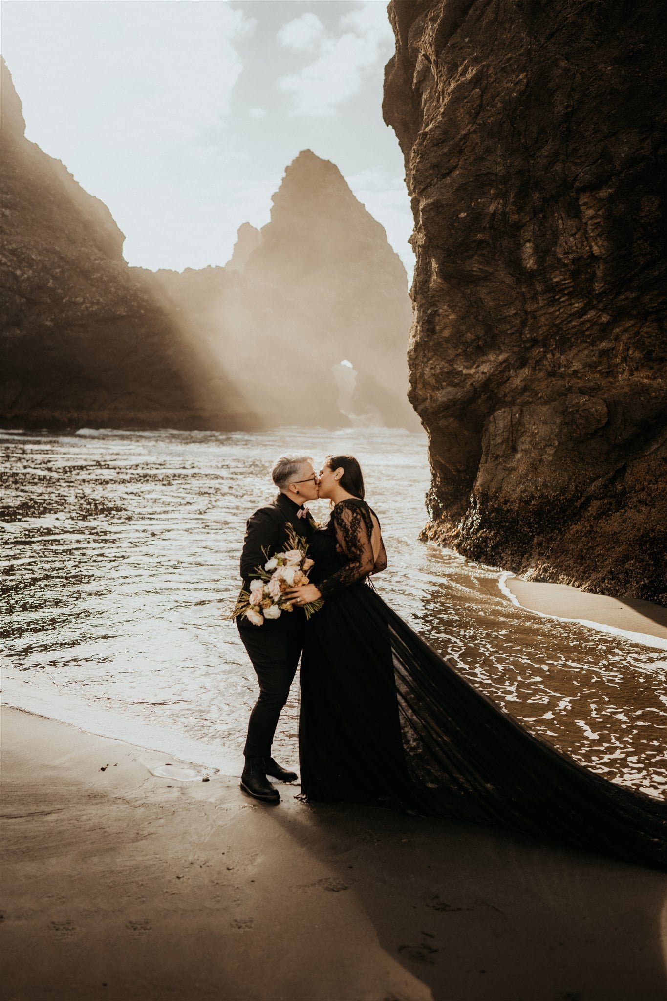 Brides kiss on the beach while wearing all black wedding outfits