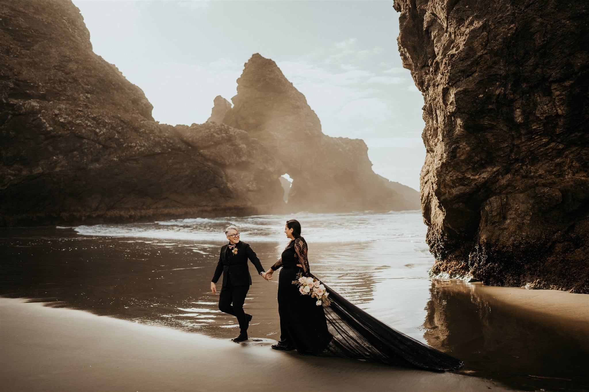 Brides hold hands and walk across the beach during their Oregon Coast elopement