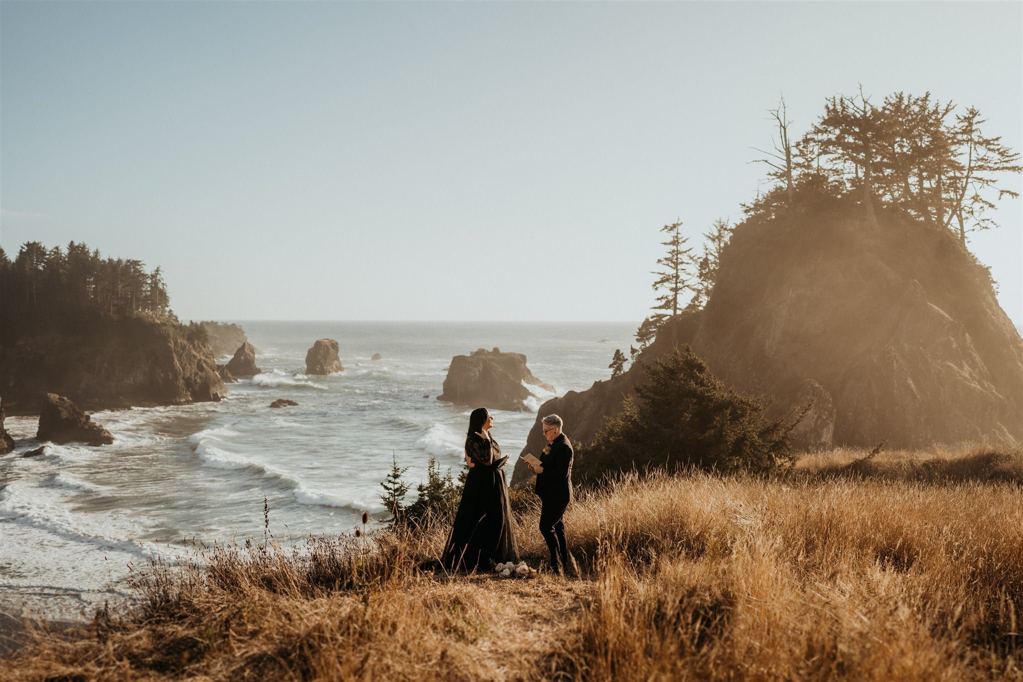 Two brides exchange vows while standing on a hill overlooking the ocean during their Pacific Northwest elopement