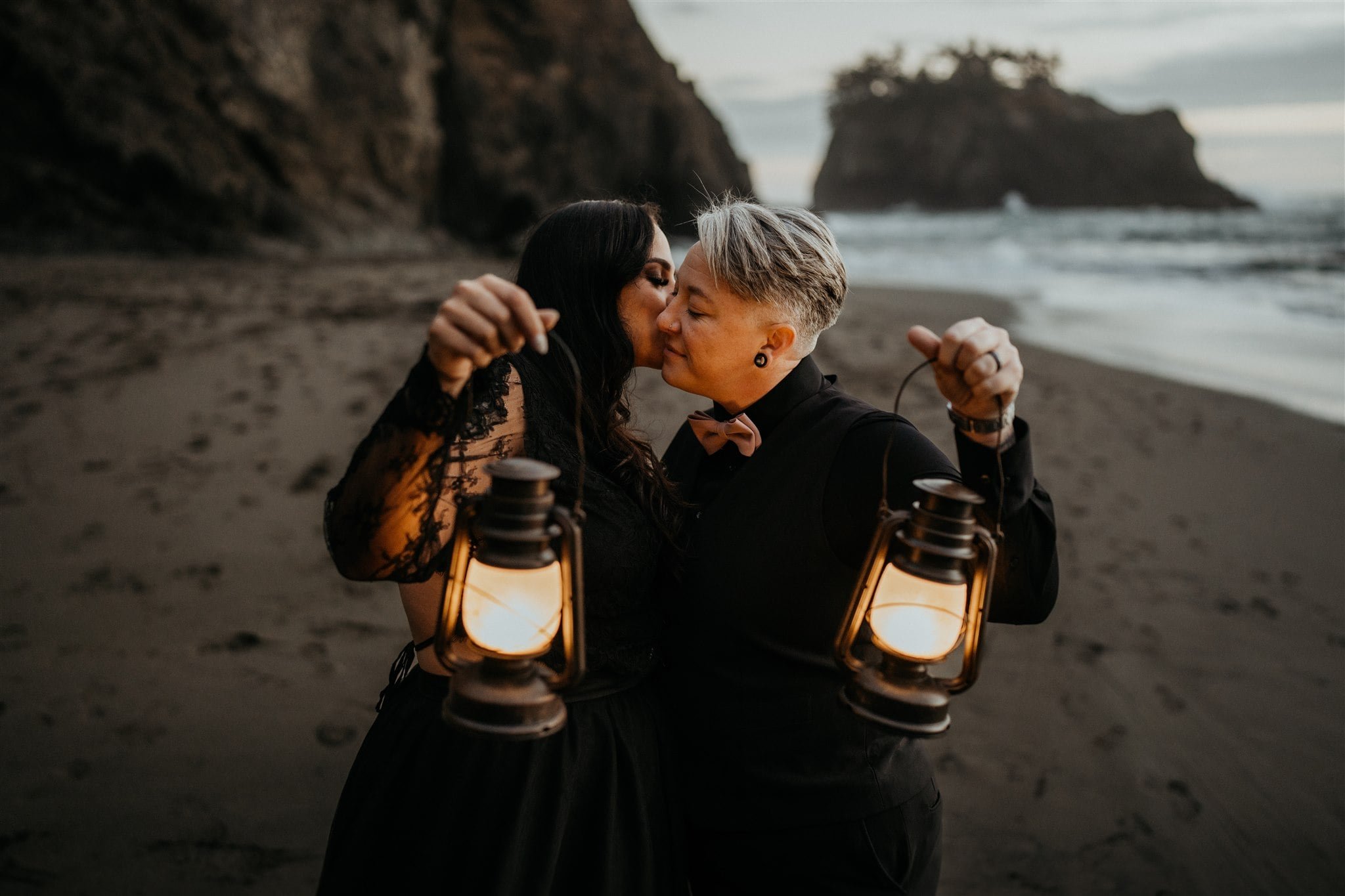 Two brides kiss while holding lanterns on the beach during their Oregon Coast elopement