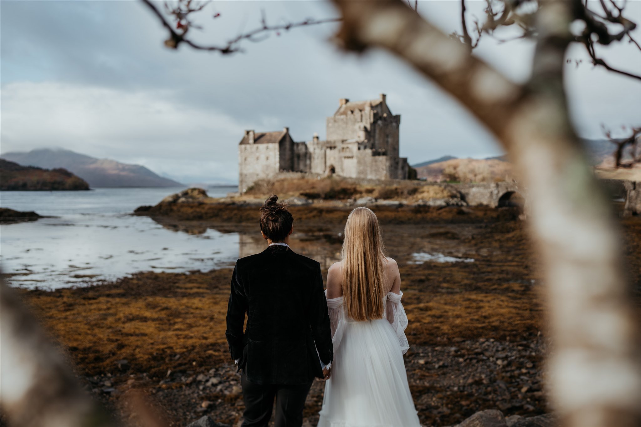 Brides hold hands while looking out at a castle on the Isle of Skye