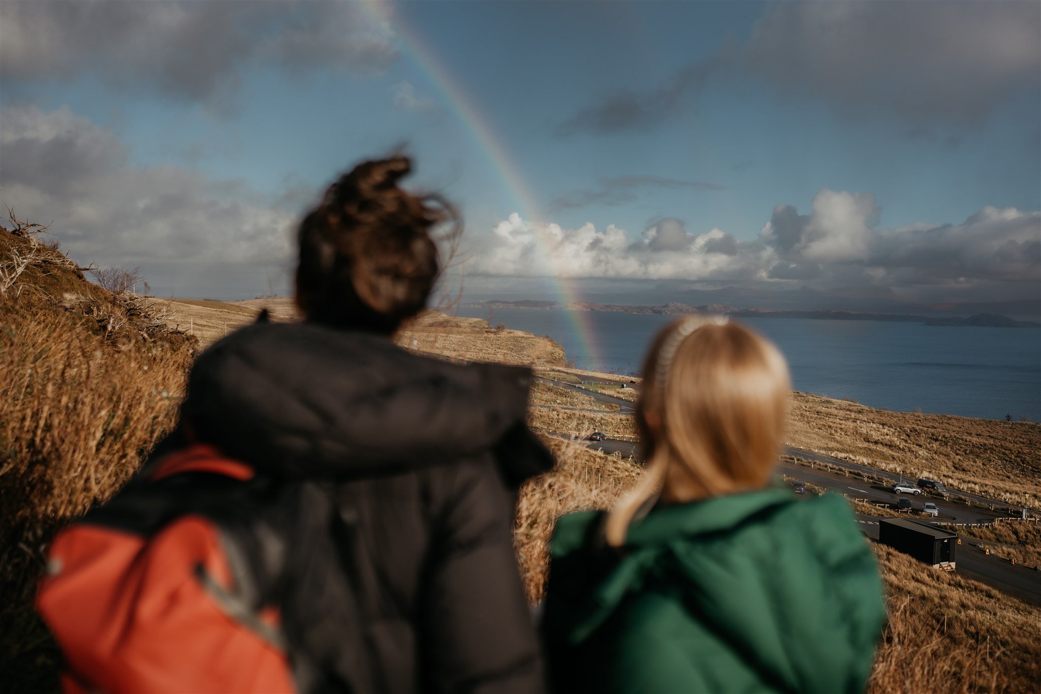 Brides look at the rainbow in the sky while hiking to their Isle of Skye elopement ceremony